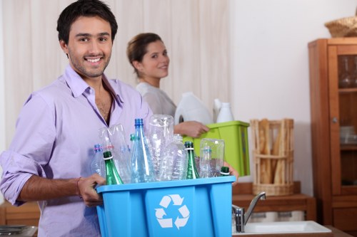 In a domestic kitchen setting, a young man with dark hair and a light purple shirt is smiling and holding a blue recycling bin filled with clear glass bottles and jars, some with visible metal caps. Behind him, a woman with long brown hair, dressed casually in light clothing, faces slightly to the side next to a green storage box, with her chair and a wooden cabinet visible in the background. The kitchen features light-colored curtains, a wooden cupboard, and a countertop with a sink, suggesting an organized, clean environment. The scene emphasizes responsible waste sorting and collection, aligning with rubbish removal services offered by Commercial Waste Removal Blackwall, highlighting the importance of proper glass recycling within local waste management efforts in London’s Blackwall area. Natural daylight illuminates the scene, emphasizing the textures and transparency of the glass bottles. The image conveys a practical and professional approach to domestic waste disposal and recycling, supporting local rubbish removal solutions.