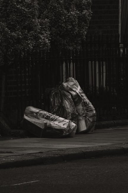Two large black garbage bags filled with waste materials rest on a paved area beside a black metal fence and a dark brick wall, adjacent to a curb on a city street or pavement. The bags appear to be m