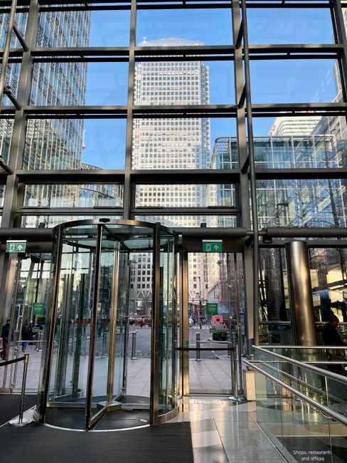 Inside a modern commercial building with large floor-to-ceiling glass windows, the image shows an entrance revolving door constructed with transparent glass panels and metallic frames, leading onto a