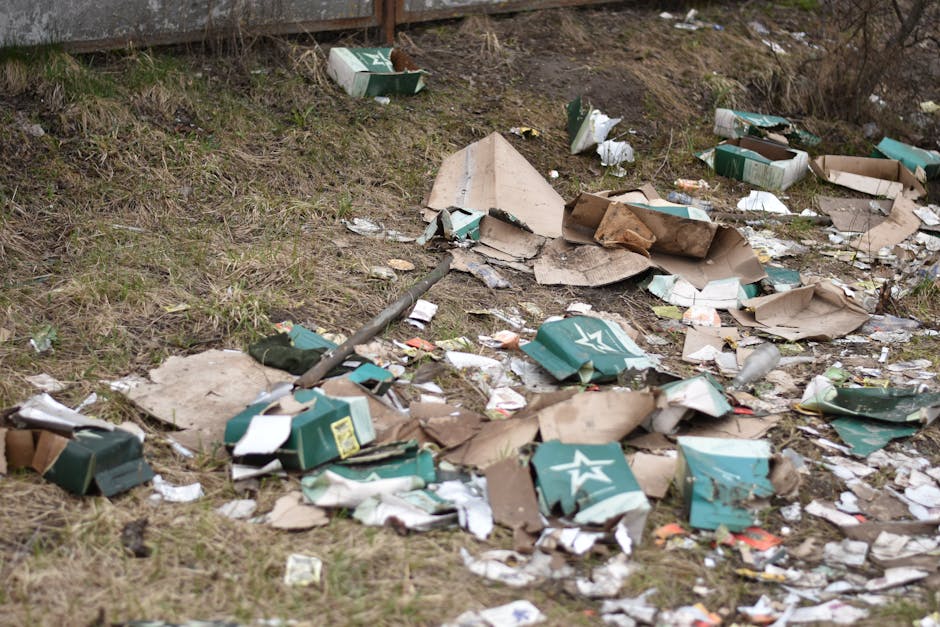An outdoor scene showing scattered cardboard boxes, some crushed or torn, along with discarded paper and plastic packaging strewn across a grassy area near a sloped dirt embankment. A metal pole or ro